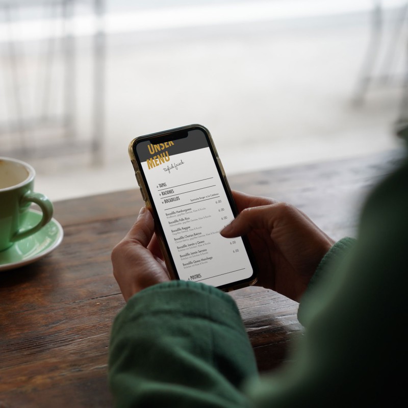 A table with a green coffee cup. In the foreground, two hands holding a smartphone and scrolling through a digital menu.