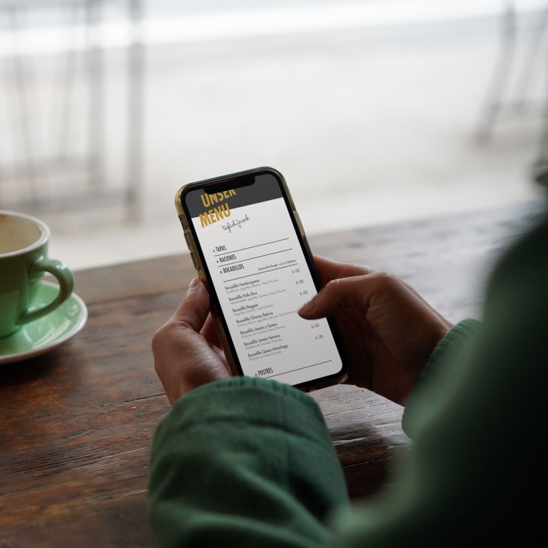 A table with a green coffee cup. In the foreground, two hands holding a smartphone and scrolling through a digital menu.